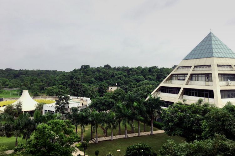 Campus der Jagran Lakecity University (JLU) im indischen Bhopal. Am rechten Bildrand ist ein pyramidenförmiges Gebäude zu sehen. Rundherum stehen Bäume, darunter einige Palmen. Zwischen den Bäumen sind Wege und eine Sportanlage zu sehen.  
