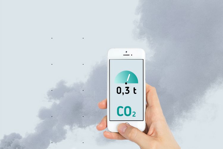 Eine Hand hält ein Smartphone ins Bild. Auf dem Bildschirm sind ein Tachometer und die Aufschrift "0,3t CO2" abgebildet. Im Hintergrund ist eine Rauchwolke zu sehen.