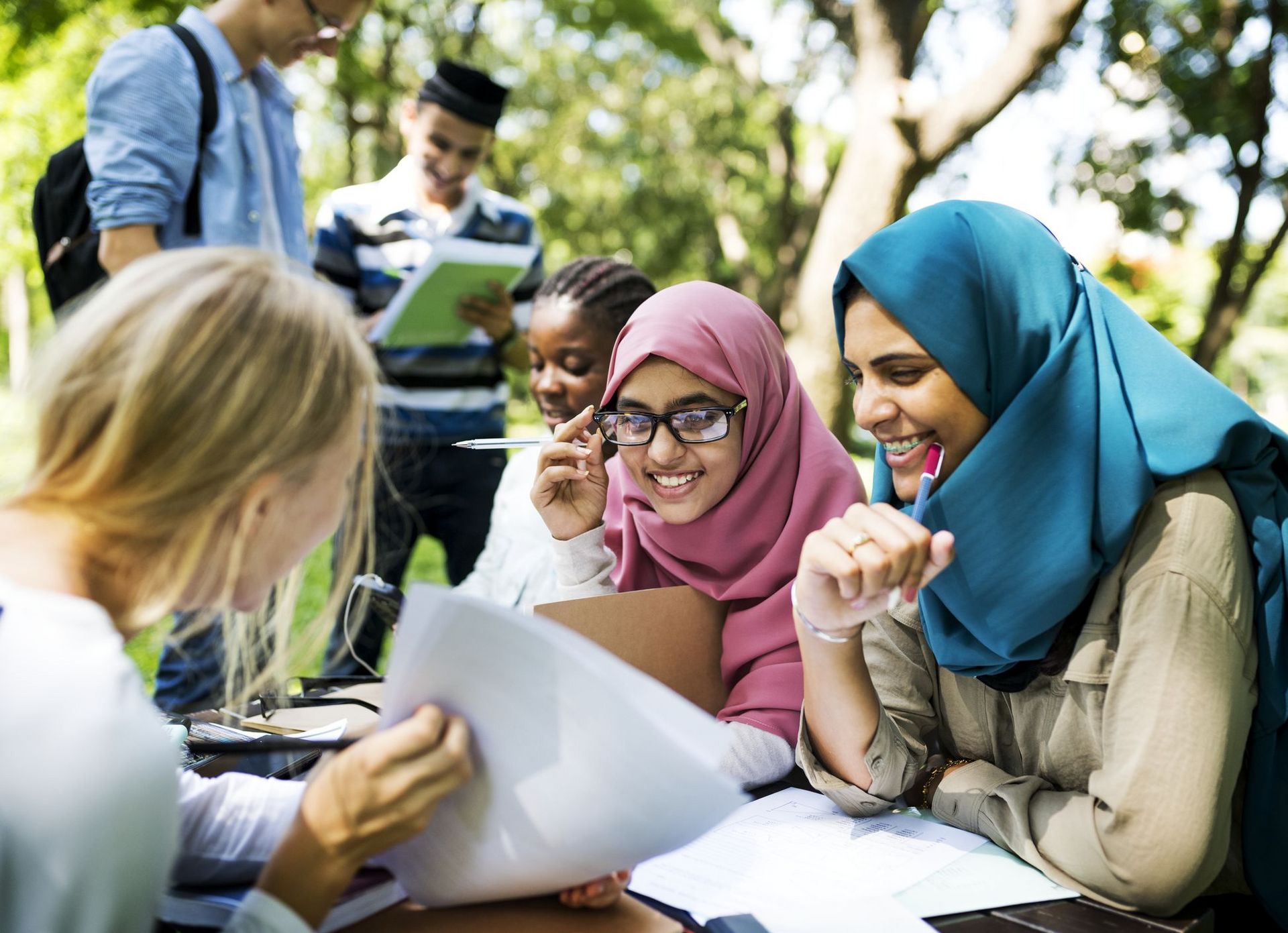 Drei weibliche Studierende (unterschiedliche Nationalitäten) in einem Campusgarten, die gemeinsam lernen und sich Skripte ansehen. Im Hintergrund stehen zwei weitere Studierende mit Rucksack und Lernmaterialien in der Hand.