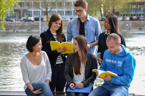 A group of students sits in conversation by the river Rhein. Some of them are holding books in their hands.