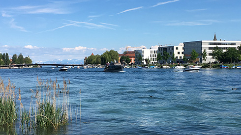 Blick über einen Fluss aufs andere Ufer, an dem größere Gebäudekomplexe stehen, blauer Himmel, im Hintergrund Berge