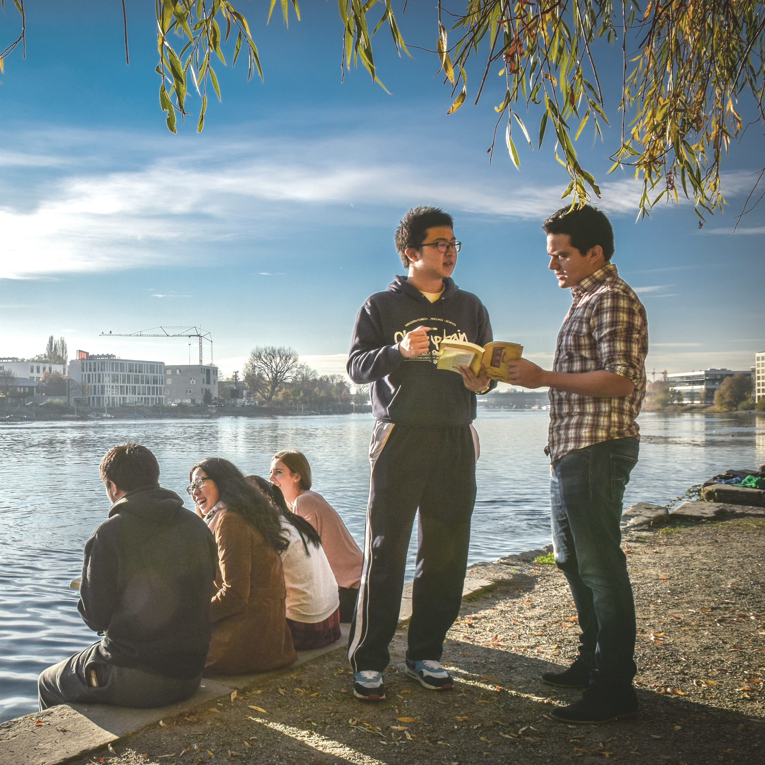 Eine Gruppe asiatischer Studierender am Seerhein, die sich unterhalten. Gegenüber des Ufers ist ein Gebäude der HTWG zu sehen.