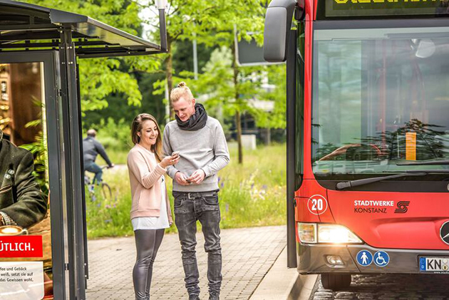Das Bild zeigt zwei Studierende an einer Bushaltestelle. Ein Bus hat gerade angehalten.