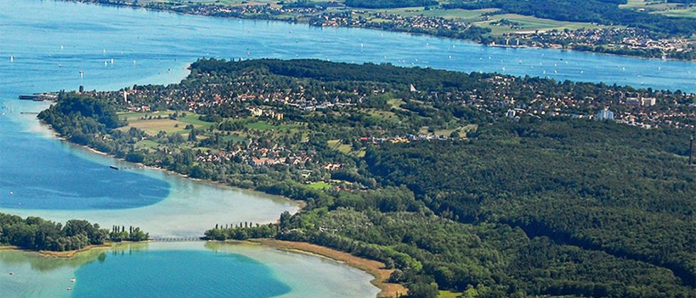 Luftaufnahme von Konstanz und dem Bodensee. Im Vordergrund liegt die Insel Mainau. Im Hintergrund sind die Alpen zu sehen.