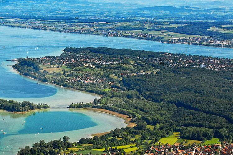 Luftaufnahme von Konstanz und dem Bodensee. Im Vordergrund liegt die Insel Mainau. Im Hintergrund sind die Alpen zu sehen.