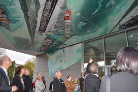 Eine Gruppe Menschen steht vor dem Eingang eines Gebäudes. Die Decke der großzügigen Überdachung des Eingangs ziert ein Kunstwerk. Zu sehen sind Formen auf einem unruhigen blauen Hintergrund, die Containerschiffen ähneln. Die Menschen blicken hinauf, einer zeigt nach oben.