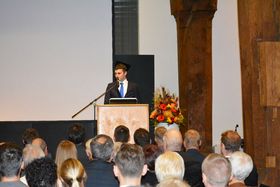Ein junger Mann im Anzug steht hinter einem Rednerpult auf einer festlich dekorierten Bühne und spricht. Vor der Bühne sitzen Zuschauer.