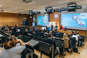 Das Bild zeigt einen Vorlesungssaal mit Publikum. An der Wand vorne befinden sich zwei Powerpointpräsentationen mit blauem Hintergrund.