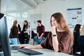 Eine junge Frau sitzt an einem Computerarbeitsplatz in einem hellen Hochschulraum, die Hand nachdenklich ans Kinn gestützt. Im Hintergrund stehen und sitzen weitere Studierende in Gruppen.
