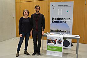 Eine junge Frau und ein junger Mann posieren neben einem Infotisch fürs Foto. An der Tischkante hängt ein Plakat mit dem Titel "Psychisch fit studieren".