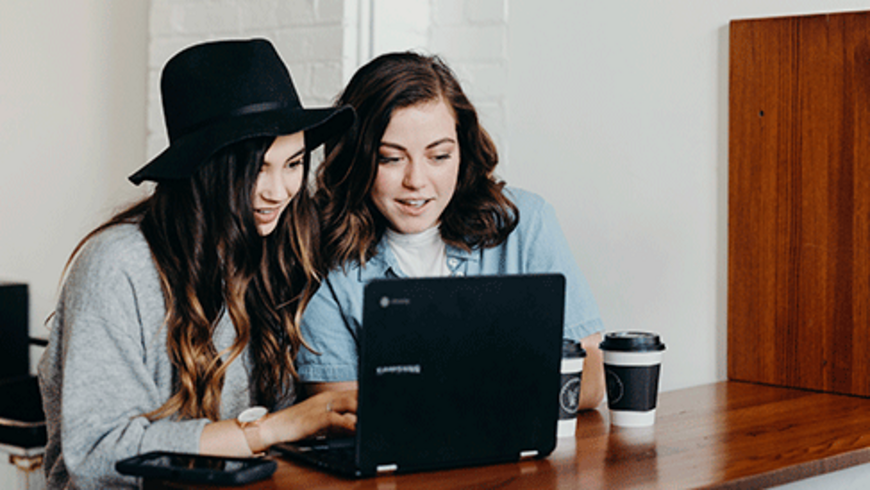 Zwei junge Frauen sitzen vor einem Laptop und unterhalten sich