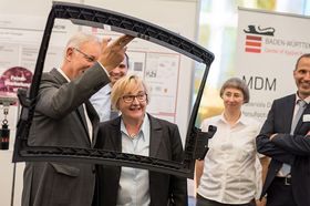 Wissenschaftsministerin Theresia Bauer posiert mit einem Mann vor einem Messestand fürs Foto. Der Mann hält einen Rahmen aus Plastik, möglicherweise ein Bauteil einer Auto-Frontscheibe. Die Ministerin und er schauen hindurch.