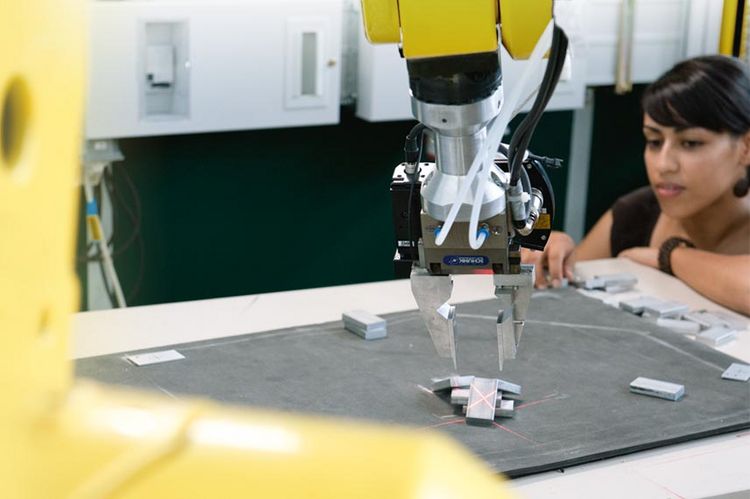 Detailansicht eines gelben Mehrachsroboters, der mit dem Greifer ein Metallstück auf einer grauen Platte anfährt. Im HIntergrund ist der Oberkörper einer Studentin zu sehen, die den Greifarm beobachtet. 