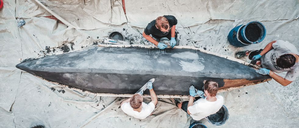 Blick von oben auf die Unterseite eines Kanus aus Beton. Um das Kanu stehen fünf Studierende, die an dem Rumpf arbeiten.