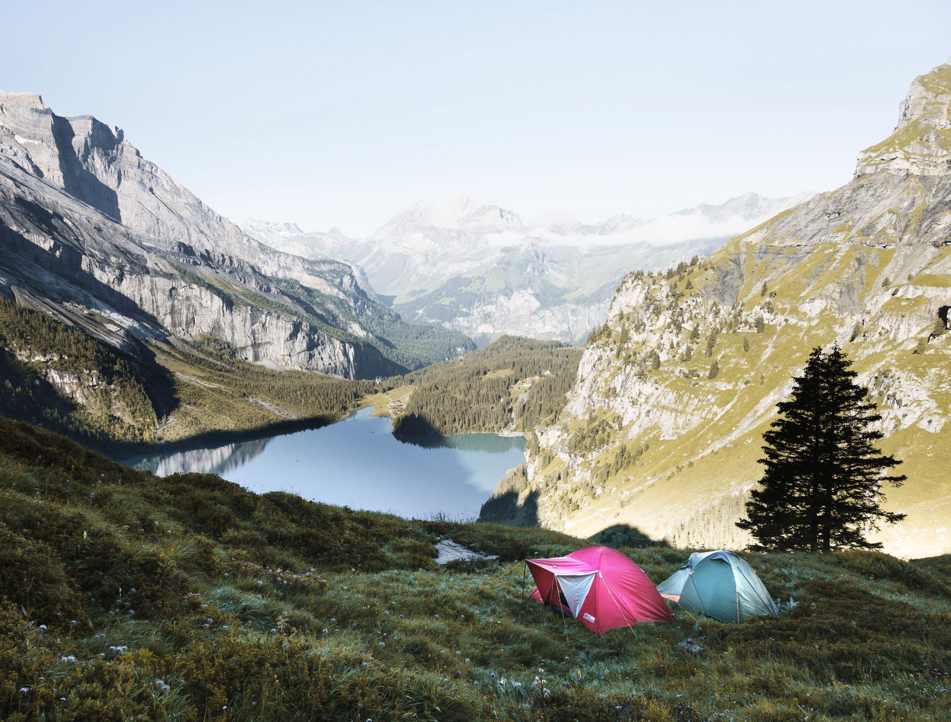 Ein rotes und ein blaues Zelt stehen auf einer Bergwiese in den Alpen. Im Hintergrund sind ein Tal mit einem See und die Berge zu sehen.