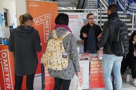 Drei Studierende sind von hinten zu sehen. Sie stehen vor einem Informationsstand von Kilometer1, der mit Postern und einem Roll-Up auf die Gründerinitiative aufmerksam macht.