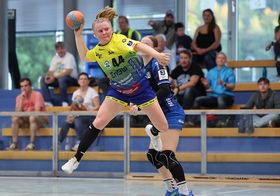 Das Bild zeigt eine Handballspielerin in Aktion. Sie springt hoch, hält in der linken Hand den Ball, ihr Gesicht ist konzentriert. Hinter ihr versucht eine Spielerin der gegnerischen Mannschaft ihr den Bald abzunehmen