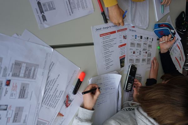 Ein Mädchen beugt sich über einen Schreibtisch mit vielen bedruckten Blättern und einem Taschenrechner.