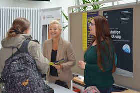 Zwei Frauen stehen hinter einem Tisch vor einer Pinnwand mit Plakaten. Sie sind im Gespräch mit einer Studentin, die vor dem Tisch steht.