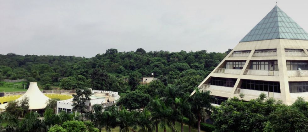 Campus der Jagran Lakecity University (JLU) im indischen Bhopal. Am rechten Bildrand ist ein pyramidenförmiges Gebäude zu sehen. Rundherum stehen Bäume, darunter einige Palmen. Zwischen den Bäumen sind Wege und eine Sportanlage zu sehen.  