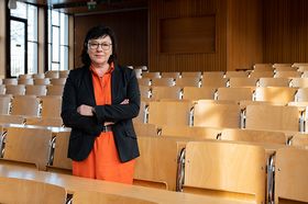 Sabine Rein steht mit verschränkten Armen in einem Hörsaal mit Holzstühlen. Sie trägt ein organgefarbenes Kleid und einen schwarzen Blazer. Ihr Blick ist freundlich und konzentriert.