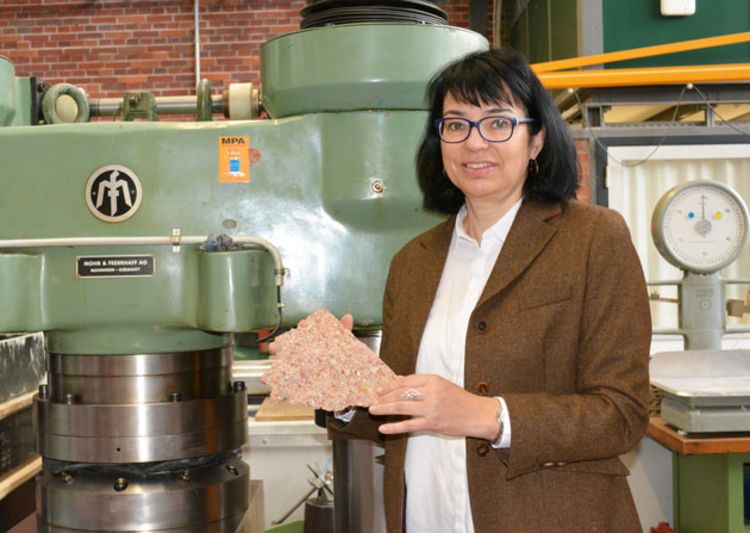 Professorin Sylvia Stürmer vor einer Maschine. Sie hält ein Stück Beton in der Hand.