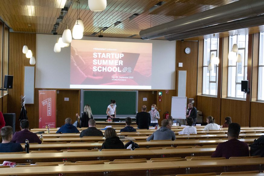 15 junge Menschen sitzen in großem Abstand voneinander in der Aula der HTWG. Auf einer Leinwand über der Tafel ist eine Präsentationsfolie mit der Aufschrift "Startup Summer School #2" zu sehen. Vor der Tafel stehen zwei junge Frauen mit Mund-Nasen-Schutz.