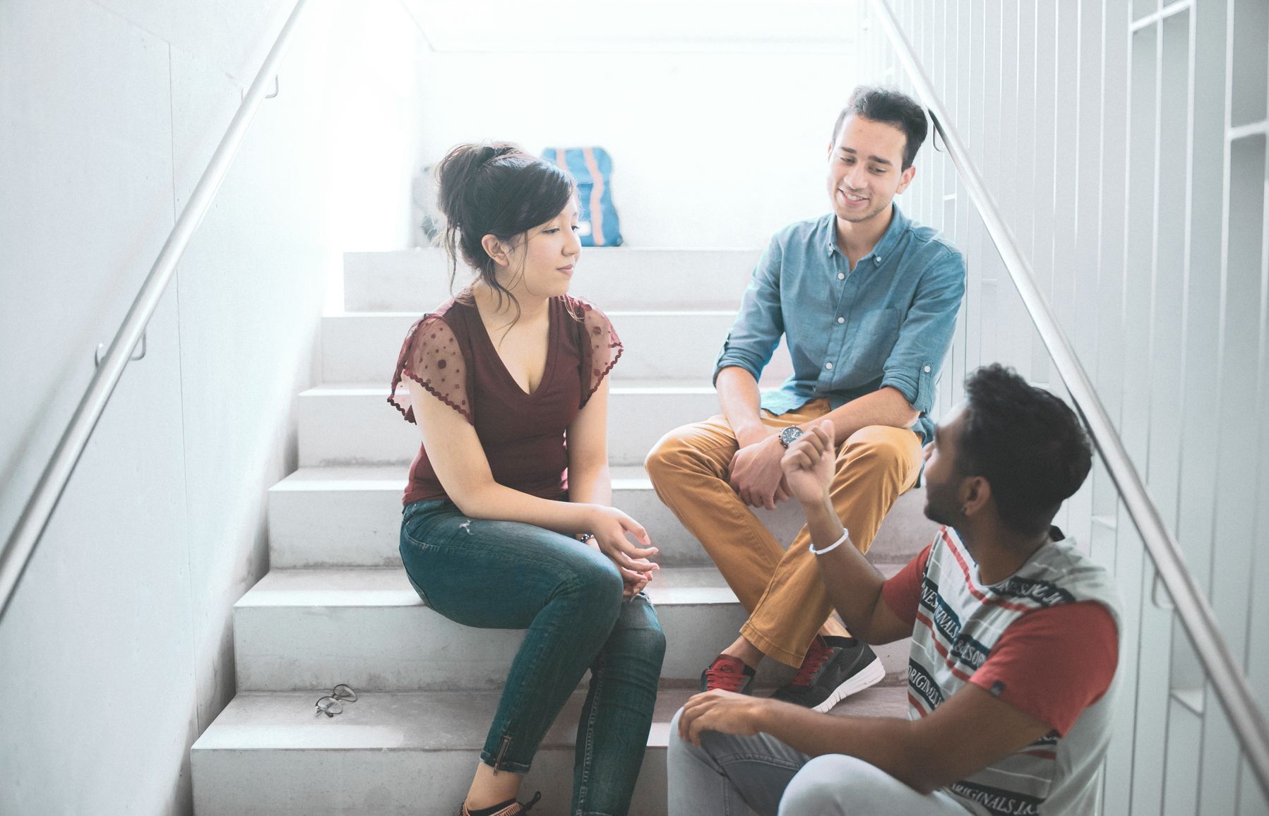 Drei Studierende sitzen auf Treppen der Hochschule und unterhalten sich Drei Studierende sitzen auf Treppen der Hochschule und unterhalten sich