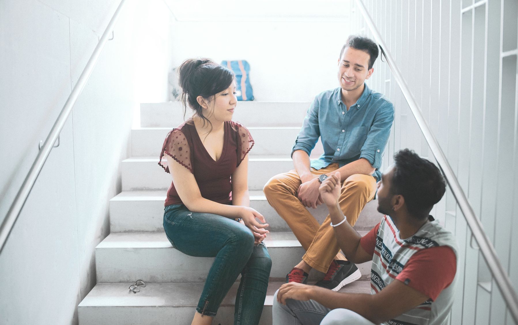 Drei Studierende sitzen auf Treppen der Hochschule und unterhalten sich Drei Studierende sitzen auf Treppen der Hochschule und unterhalten sich