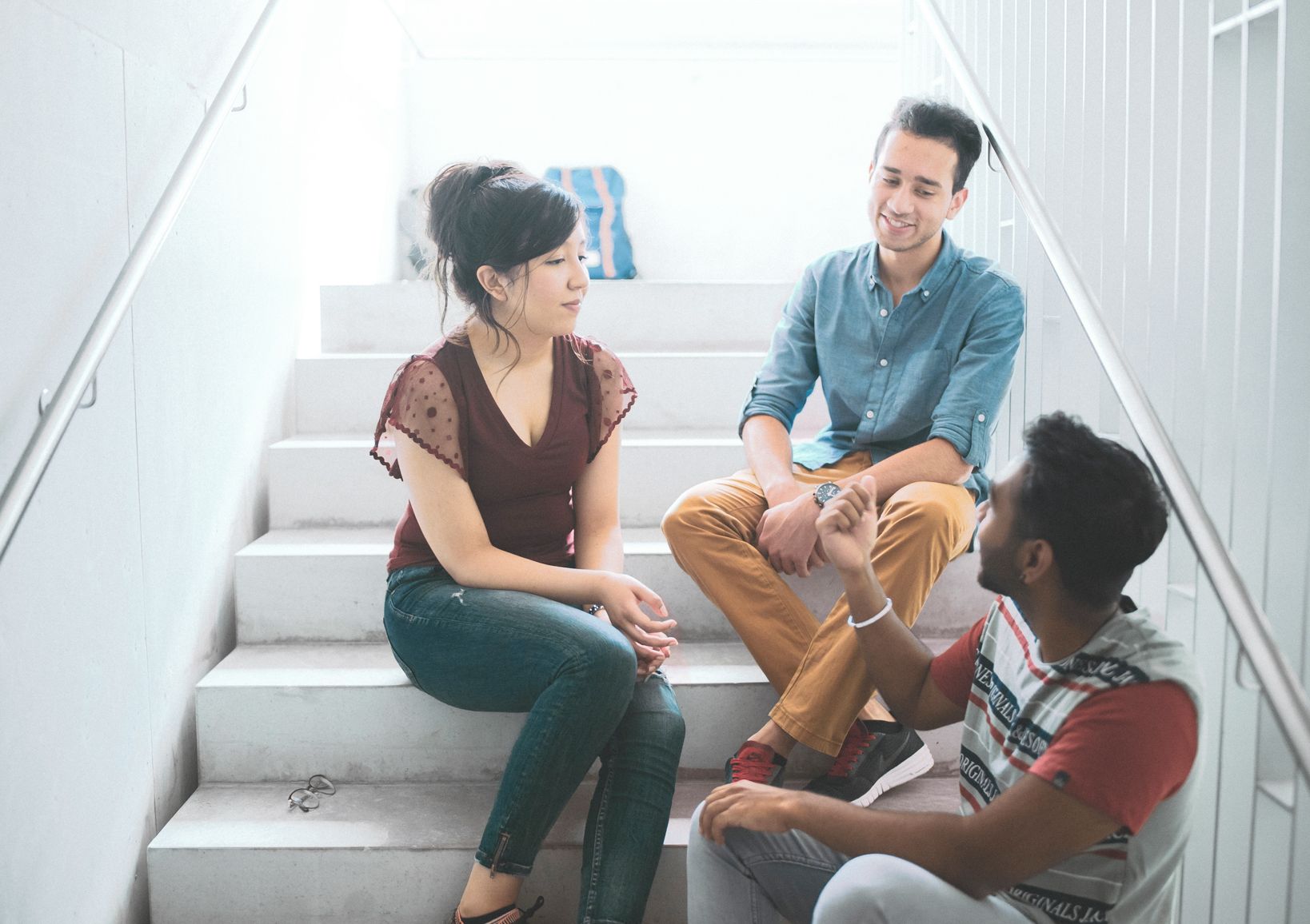 Drei Studierende sitzen auf Treppen der Hochschule und unterhalten sich Drei Studierende sitzen auf Treppen der Hochschule und unterhalten sich