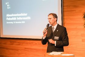 Person mit Mikrofon vor Leinwand, auf der das HTWG-IN-Logo abgebildet ist sowie folgender Text: "Absolventenfeier Fakultät Informatik, Donnerstag, den 13. November 2025"