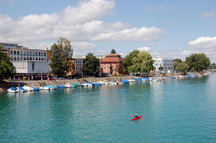 Ein rotes Kanu fährt mitten auf dem Konstanzer Seerhein. Im Hintergrund ist der Campus der HTWG zu sehen.