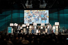 Studierende stehen bei der 120-Jahr-Feier der HTWG auf der Bühne des Bodenseeforums und halten Schilder hoch, die zusammen den Satz ‚Für die Zukunft. Seit 1906.' ergeben. Im Hintergrund läuft ein Gruppenfoto des Publikums auf der Leinwand, davor sitzen die Gäste im dunklen Saal.