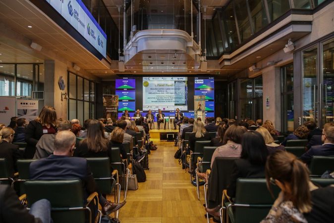 Das Veranstaltungsbild zeigt das Auditorium und am Ende das Raums die Podiumsdiskussion mit den zugehörigen Präsentationsfolien.