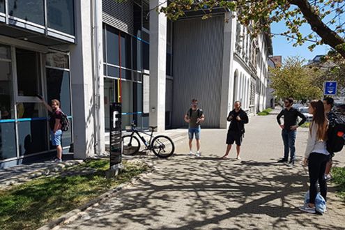 Studierende stehen vor dem F-Gebäude und warten auf die Ausgabe von Material, was Coronabedingt durch ein Fenster herausgericht werden muss.