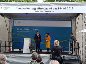 Eine Frau in einem braunen Ledermantel hält ein Handmikrofon in der Hand und blickt nach links auf einen Mann im Anzug, der ebenfalls ein Handmikrofon in der Hand hält. Die beiden stehen auf einer Bühne, hinter der Bühne hängt eine blaue Leinwand , auf der steht: Transferinitiative.