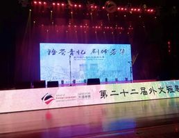 Ein dunkler Veranstaltungssaal  der School of Foreign Languages der Qingdao Universität. Der Beamer wirft ein Bild mit Informationen auf Chinesisch an die Wand. Rundherum ist es dunkel. Vor dem Beamer steht ein langer Tisch mit leeren Stühlen für die Präsentierenden. An der Decke leuchten pinke Lichter. Der Boden vor dem langen Tisch spiegelt das Licht. 