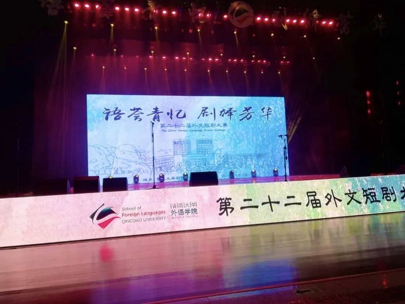 Ein dunkler Veranstaltungssaal  der School of Foreign Languages der Qingdao Universität. Der Beamer wirft ein Bild mit Informationen auf Chinesisch an die Wand. Rundherum ist es dunkel. Vor dem Beamer steht ein langer Tisch mit leeren Stühlen für die Präsentierenden. An der Decke leuchten pinke Lichter. Der Boden vor dem langen Tisch spiegelt das Licht. 