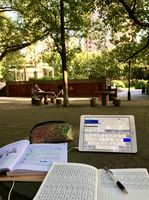 Auf einem Tisch im Freien in einem Park auf dem Campus liegen ein Chinesisch-Lernbuch, ein Tablet, welches als Wörterbuch verwendet wird, ein Übungsheft zum Schreiben von Schriftzeichen und ein glitzerndes Stifteetui. Im Hintergrund sieht man zwei Tische neben einem Baum. An einem der beiden Tische in der Ferne sitzt ein chinesischer Mann. 