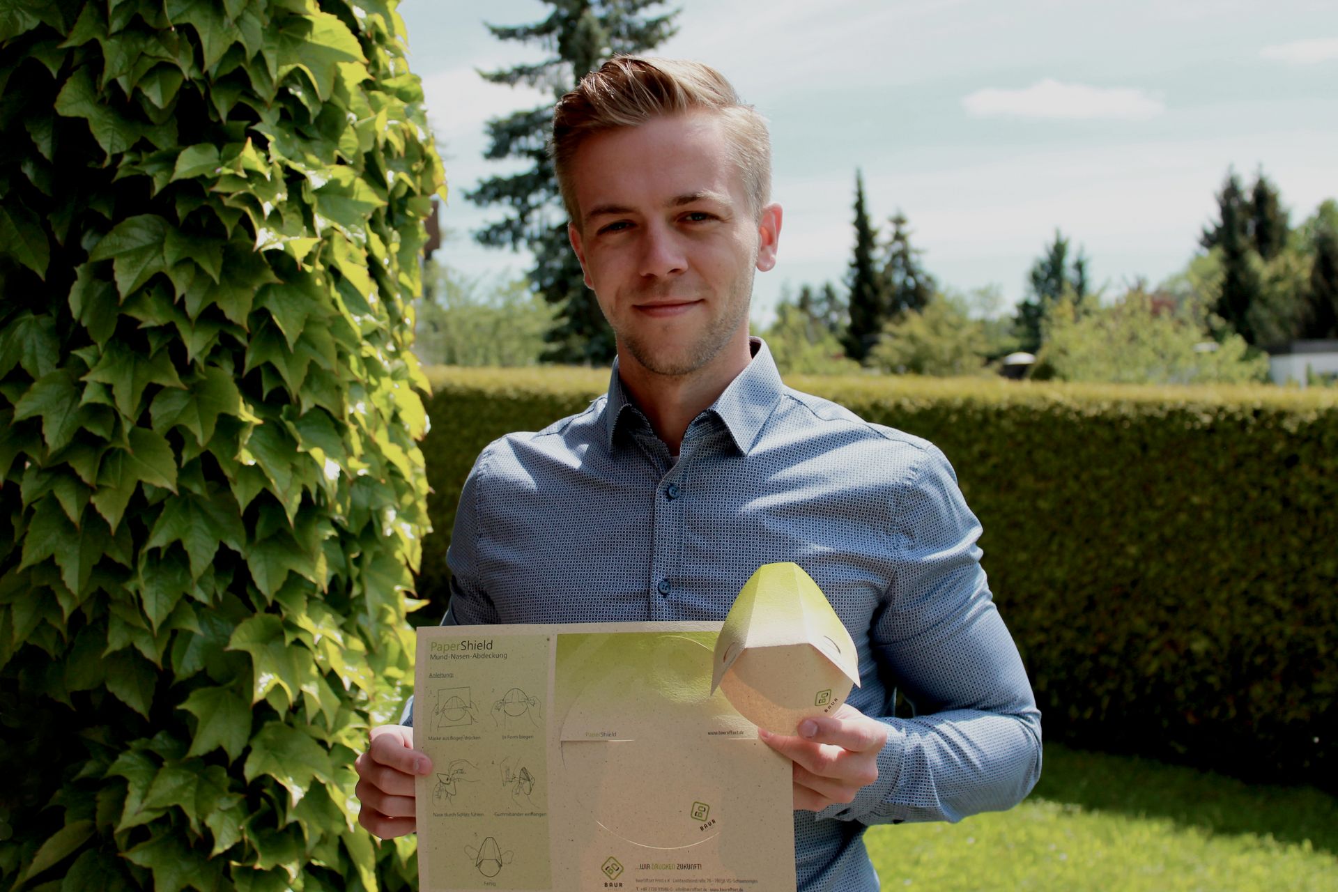 Tim Mauch steht in einem Garten und hält einen Papierbogen mit Maske und Nutzungsanleitung sowie eine fertig geformte Maske in den Händen.