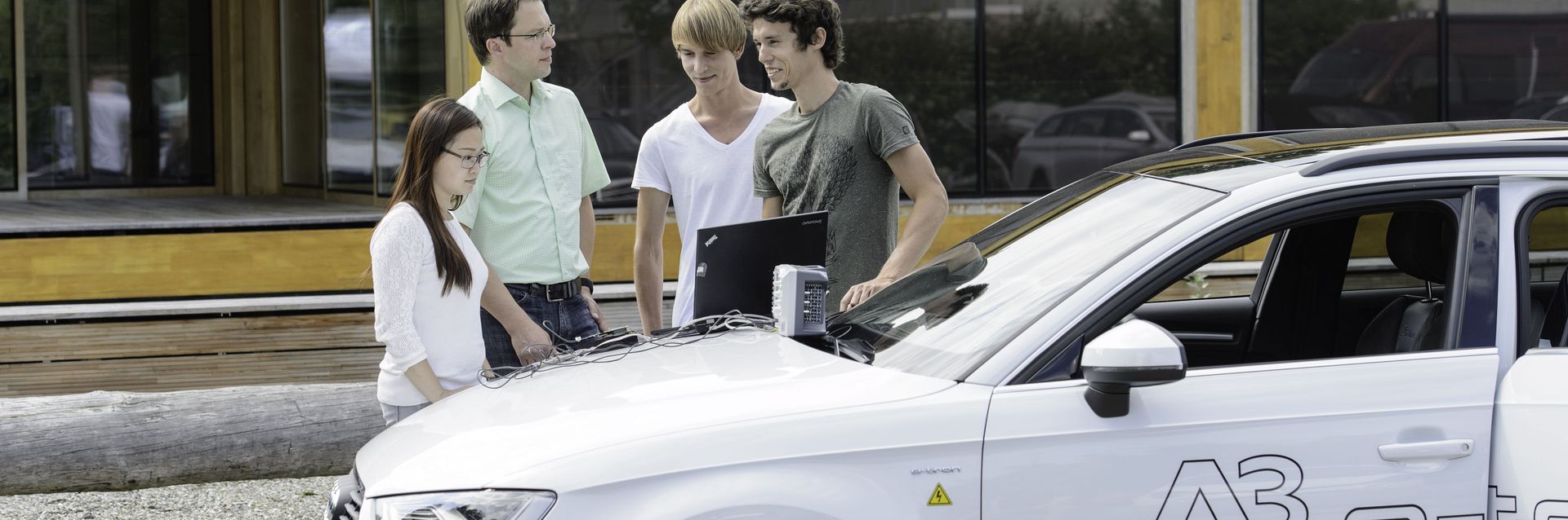Professor Lang steht mit einigen Studierenden vor einem Audi etron, auf dessen Motorhaube ein Laptop zur Steuerung und Bearbeitung liegt. 