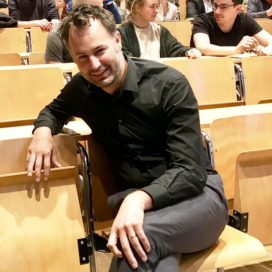 Portrait von Dan Shürich, der in einem Vorlesungssaal auf einem Holz-Klappstuhl sitzt.