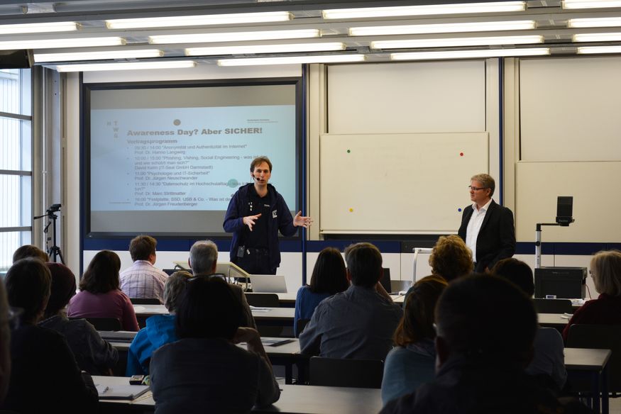 Prof. Dr. Hanno Langweg (links) und Prof. Dr. Jürgen Freudenberger (rechts) stehen in einem Hörsaal und blicken ins Publikum.