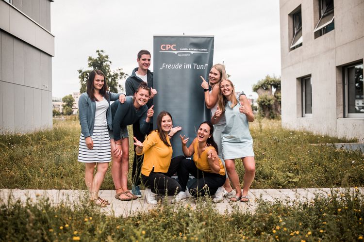 Sech Studentinnen und ein Student stehen lachend um ein Roll-up und blicken in die Kamera. Auf dem Roll-up steht: CPC - Freude im Tun.