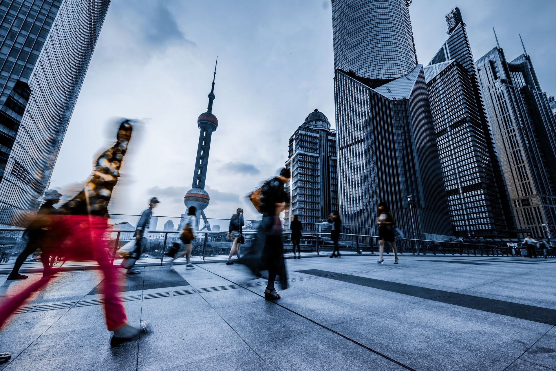 Firmengebäude (Skyline) einer größeren chinesischen Stadt. Im Vordergrund laufen verschiedene Menschen über einen Platz, die jedoch verschwommen dargestellt sind, damit das Bild dynamisch wirkt und ein hektisches Arbeitsleben widerspiegelt.