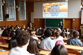 Hinterköpfe junger Menschen in einer Aula, vorne spricht ein Mann im Anzug.