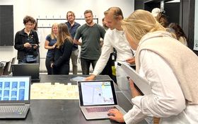 DasFoto zeigt eine Gruppe von Menschen, die um einen Tisch stehen. Eine Frau und ein Mann schauen au einen Laptop