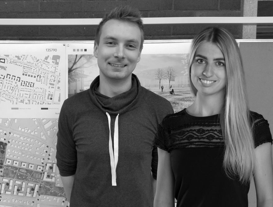 Das schwarz-weiß Foto zeigt Adrian Buck und Denice Müller vor einer Stellwand mit Architekturplänen.