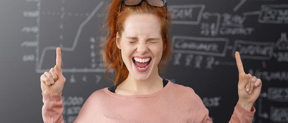 Eine junge Frau in einem rosa Shirt steht vor einer Schiefertafel, auf der mathematische Formeln mit Kreide geschrieben stehen. Die junge Frau hat die beiden Arme erhoben und an beiden Fäusten jeweils den Zeigefinger ausgestreckt. Sie hat die Augen zusammengekniffen und den Mund offen.