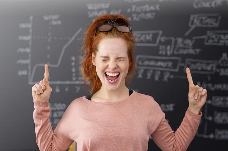 Eine junge Frau in einem rosa Shirt steht vor einer Schiefertafel, auf der mathematische Formeln mit Kreide geschrieben stehen. Die junge Frau hat die beiden Arme erhoben und an beiden Fäusten jeweils den Zeigefinger ausgestreckt. Sie hat die Augen zusammengekniffen und den Mund offen.